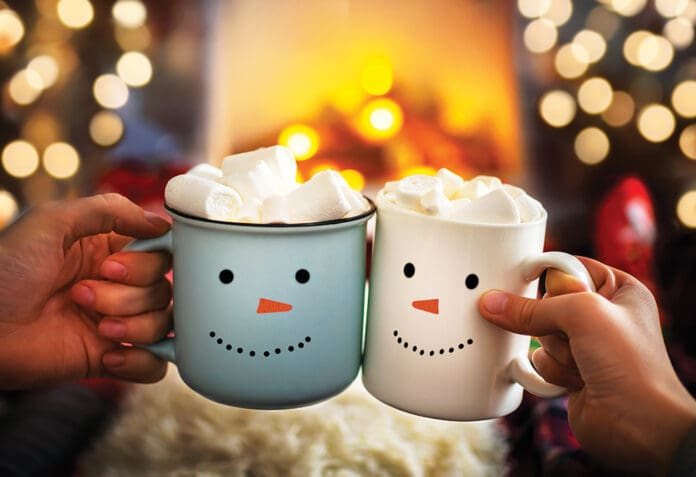 Close-up of two hands with snowman face on cup of hot cocoa with marshmallows. Mom and child relaxing together on a cozy winter evening by fireplace. Enjoy Christmas holidays, happy moments at home.